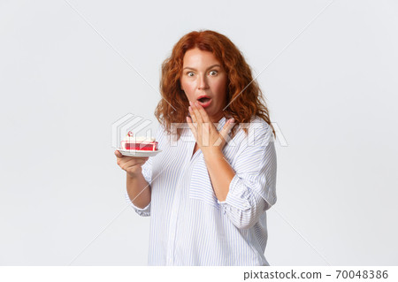 Holidays, emotions and lifestyle concept. Shocked and concerned middle-aged redhead woman open mouth, gasping and looking worried while holding cake, hear how much calories it has 70048386
