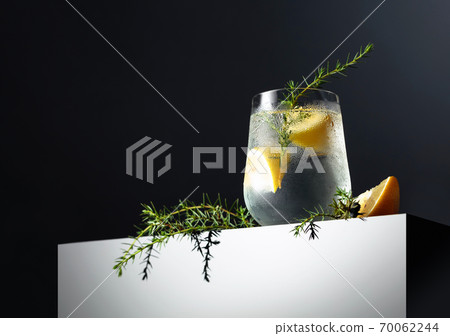 Alcohol drink (gin tonic cocktail) with lemon, juniper branch, and ice on a dark reflective background. Alcohol drink (gin tonic cocktail) with lemon, juniper branch, and ice on a dark reflective background. 70062244