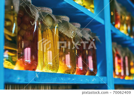 Glass jars covered with burlap fabric with different homemade canned food, preserved vegetables on wooden shelves 70062409