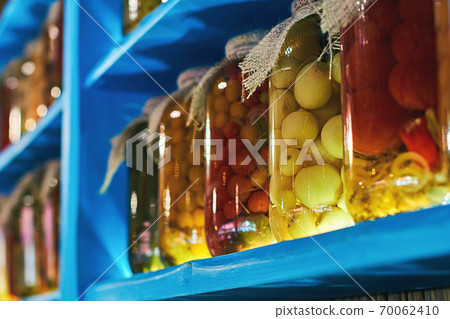 Glass jars covered with burlap fabric with different homemade canned food, preserved vegetables on wooden shelves Glass jars covered with burlap fabric with different homemade canned food, preserved vegetables on wooden shelves 70062410