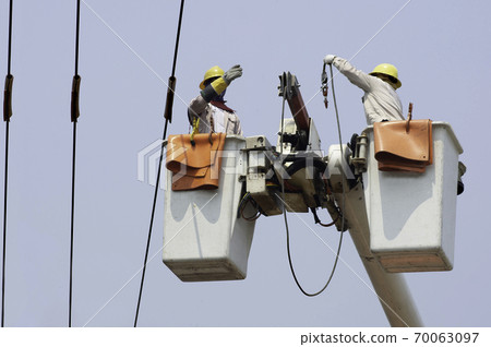 Electricians work together on the electric cable car and electric pole. To maintain the high and low voltage distribution system. They wear a helmet with PPE protection and copy space for your text. Electricians work together on the electric cable car and electric pole. To maintain the high and low voltage distribution system. They wear a helmet with PPE protection and copy space for your text. 70063097