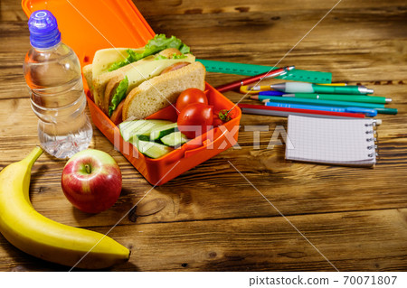 Back to school concept. School supplies, bottle of water, apple, banana and lunch box with sandwiches and fresh vegetables on a wooden desk Back to school concept. School supplies, bottle of water, apple, banana and lunch box with sandwiches and fresh vegetables on a wooden desk 70071807