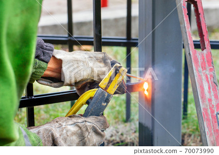 A worker welder works with metal, welds a metal fence at a construction site. 70073990