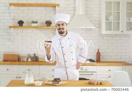 Confectioner man smiles while standing with cake in his hands at the kitchen bakery Confectioner man smiles while standing with cake in his hands at the kitchen bakery 70083446