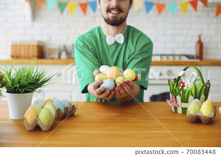 Happy Easter. A funny bearded man holds eggs in his hands and sits in the decorated room with the ears of a rabbit. Happy Easter. A funny bearded man holds eggs in his hands and sits in the decorated room with the ears of a rabbit. 70083448