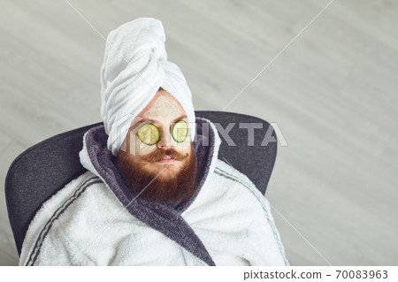 Funny fat bearded man with a cosmetic mask on his face in bathrobe towel on his head on his face resting against a gray background 70083963