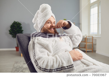 Funny strange fat bearded man with a cosmetic mask on his face in a bathrobe does spa treatments on his face rests in a room Funny strange fat bearded man with a cosmetic mask on his face in a bathrobe does spa treatments on his face rests in a room 70083967