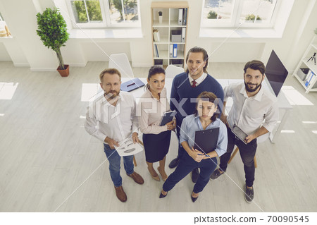 Group of smiling business people standing with documents and looking at camera ahead Group of smiling business people standing with documents and looking at camera ahead 70090545