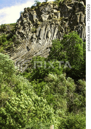 Basalt curved columns on a hill in the Garni gorge among green trees growing along the banks of the Azat river against a blue sky covered with clouds 70090620