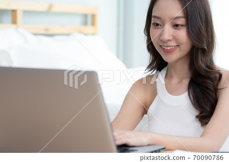 Asian woman working on a laptop sitting on the bed in the house. 70090786