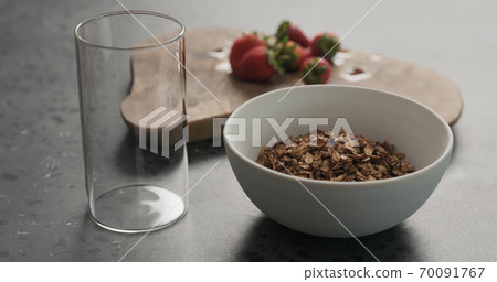 white bowl with chocolate granola, strawberries and glass for yogurt on a concrete countertop 70091767