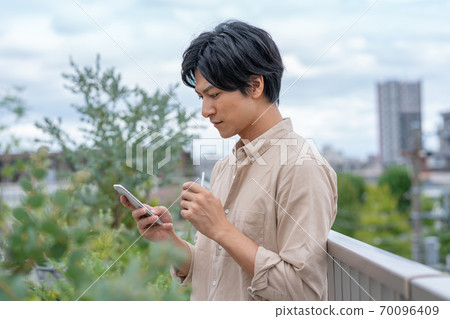 一個年輕人在建築物的屋頂上看著智能手機時抽著電子煙 70096409