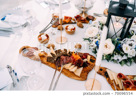 A large white candle in a black metal candlestick-lantern in a bouquet of flowers on a table with delicacies for the holiday. A large white candle in a black metal candlestick-lantern in a bouquet of flowers on a table with delicacies for the holiday. 70096554