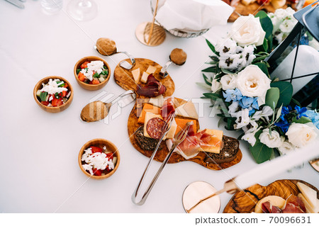 Cutting board with delicacies, kitchen tongs and spoons with a curved handle on a table with a candle in a candlestick lantern in a bouquet of flowers. 70096631