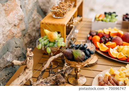 Gilded bowls with blue and white grapes, figs and pears on a wooden table with ham, cheese and fruits on a tray. 70096747