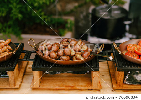 Fried halves of potatoes, in a skillet on the stove, with fried sausages and chopped tomato rings on a buffet table at the hotel. 70096834