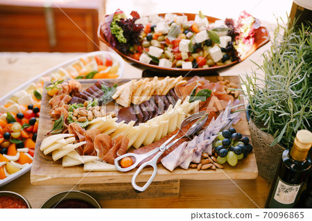 Close-up of a wooden cutting board with meat and cheese cuts, a grape cube and kitchen scissors on a background of salad and vegetables. 70096865