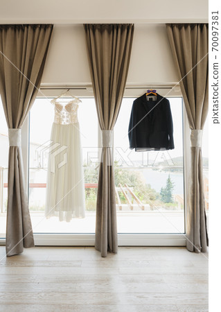 The groom's suit is black and the bride's white lace dress on hangers by the window with gray curtains. The groom's suit is black and the bride's white lace dress on hangers by the window with gray curtains. 70097381
