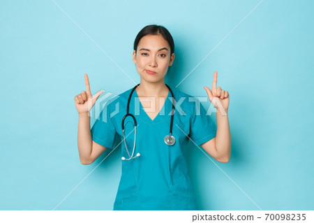 Covid-19, social distancing and coronavirus pandemic concept. Skeptical and doubtful young asian female doctor, professional physician or nurse in scrubs pointing fingers up, light blue background Covid-19, social distancing and coronavirus pandemic concept. Skeptical and doubtful young asian female doctor, professional physician or nurse in scrubs pointing fingers up, light blue background 70098235
