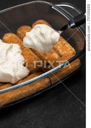 tiramisu dessert cooking - Traditional Italian Savoiardi ladyfingers Biscuits and cream in glass baking dish on dark concrete background tiramisu dessert cooking - Traditional Italian Savoiardi ladyfingers Biscuits and cream in glass baking dish on dark concrete background 70100889