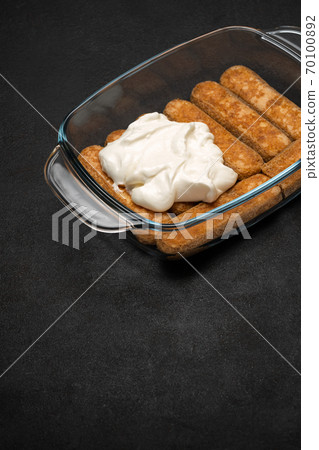 tiramisu dessert cooking - Traditional Italian Savoiardi ladyfingers Biscuits and cream in glass baking dish on dark concrete background tiramisu dessert cooking - Traditional Italian Savoiardi ladyfingers Biscuits and cream in glass baking dish on dark concrete background 70100892