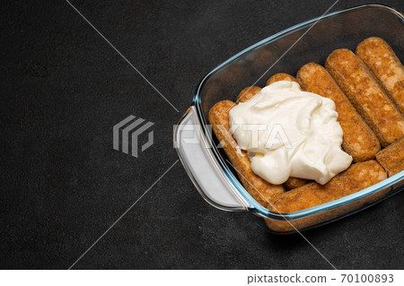 tiramisu dessert cooking - Traditional Italian Savoiardi ladyfingers Biscuits and cream in glass baking dish on dark concrete background 70100893