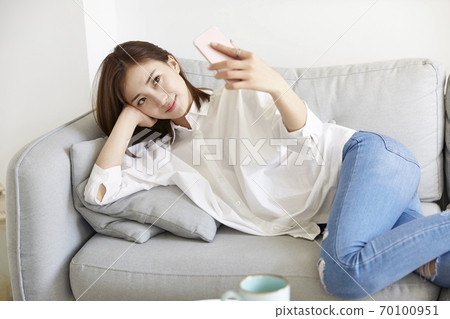young asian woman lying on sofa taking a selfie using mobile phone 70100951