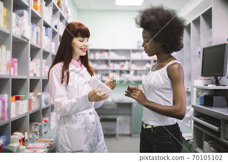 Beautiful professional female pharmacist talking with young African woman patient, while showing her two dosage forms of medicine, and explaining how to use it. Pharmaceutical care Beautiful professional female pharmacist talking with young African woman patient, while showing her two dosage forms of medicine, and explaining how to use it. Pharmaceutical care 70105102
