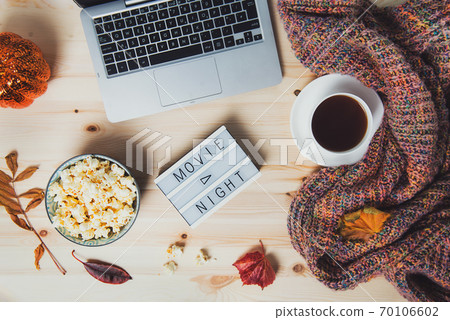 Top view Movie night concept. Flat lay composition with Movie night message on the board, laptop, popcorn bowl, decorative pumpkin, fallen leaves, a cup of tea, and warm plaid on wooden background. Top view Movie night concept. Flat lay composition with Movie night message on the board, laptop, popcorn bowl, decorative pumpkin, fallen leaves, a cup of tea, and warm plaid on wooden background. 70106602