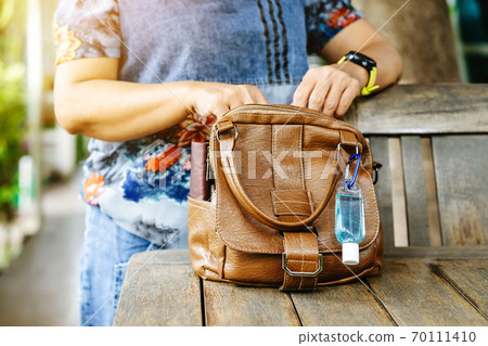 Mini portable alcohol gel bottle to kill Corona Virus(Covid-19) hang on a brown leather shoulder bag on table in garden. New normal lifestyle. Health care concept. Selective focus on alcohol gel Mini portable alcohol gel bottle to kill Corona Virus(Covid-19) hang on a brown leather shoulder bag on table in garden. New normal lifestyle. Health care concept. Selective focus on alcohol gel 70111410