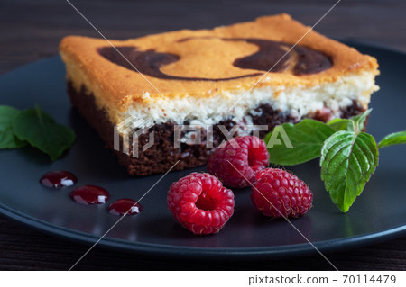 A piece of double-layer cake on a plate of chocolate brownies and curd cheesecake with raspberries. Dark background copy space 70114479
