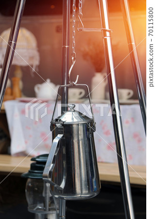 Stainless kettle hanging on the tripod and out focus cup set of tea and coffee on the table cloth Stainless kettle hanging on the tripod and out focus cup set of tea and coffee on the table cloth 70115800