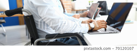 Disabled businessman sits in wheelchair in office and works at laptop Disabled businessman sits in wheelchair in office and works at laptop 70120107