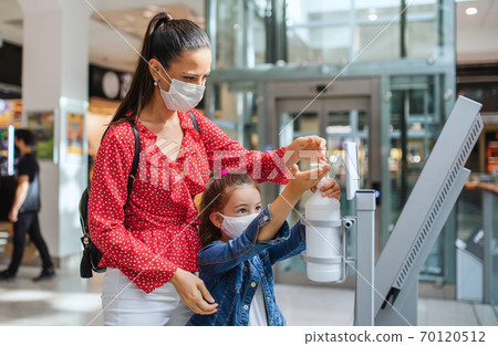 Mother and daughter with face mask disinfecting hands indoors in shopping center, coronavirus concept. 70120512