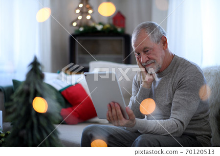 Sad and lonely senior man with tablet sitting at the table indoors at Christmas, solitude concept. 70120513