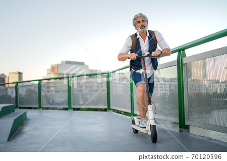 Mature man commuter with electric scooter outdoors in city, going to work. 70120596