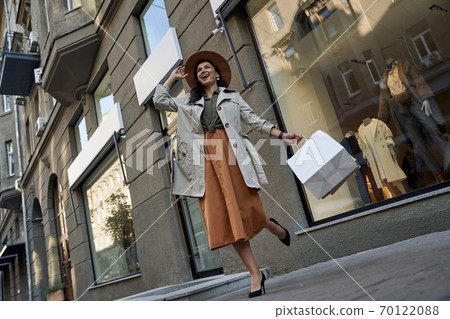 Stylish lady. Young beautiful excited woman walking city streets with shopping bags, looking aside and laughing Stylish lady. Young beautiful excited woman walking city streets with shopping bags, looking aside and laughing 70122088
