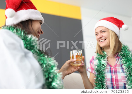Two women friends in santa claus hats hold glasses of whiskey with ice in their hands in apartment 70125835