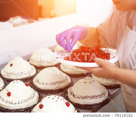 A chef decorates cakes in a confectionery factory with fresh cherry. Dessert mass production, background, industry, confectioner A chef decorates cakes in a confectionery factory with fresh cherry. Dessert mass production, background, industry, confectioner 70128182