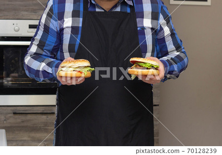 a man with a beard and in an apron holds burgers in his hands in the kitchen, background 70128239