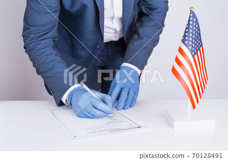 Man in protective medical gloves votes on presidential election of USA during coronavirus pandemic COVID 19. Person fills out paper ballot. Flag of the United States is on the table. General voting 70128491