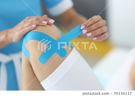 Traumatologist holds blue tape in his hand to fix patient's knee joint in clinic Traumatologist holds blue tape in his hand to fix patient's knee joint in clinic 70130117