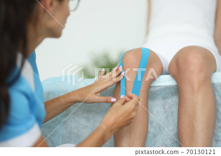 Physiotherapist sticking blue tape on patient's knee in clinic closeup 70130121