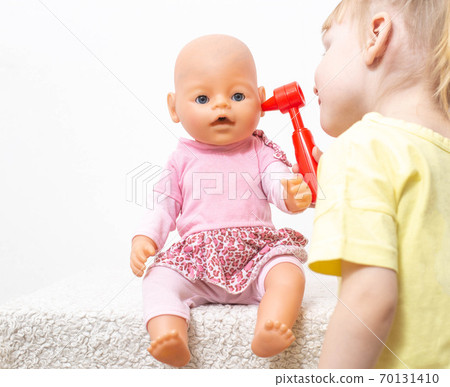 Little girl inspects the ears of her doll on a white background. The concept of pediatric otolaryngology otitis media of the ears and sulfur test in the ear, copy space 70131410