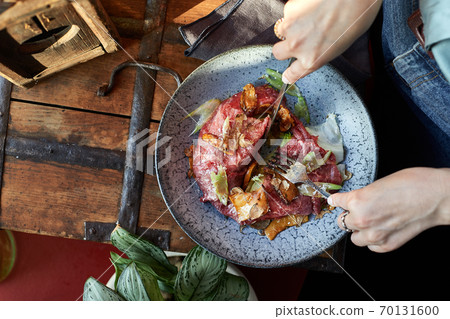 A female hand is preparing a vegetable salad with beef roast beef. A plate of salad with beef roast beef, onions, arugula, pepper and tomatoes on a black table. Top view. A female hand is preparing a vegetable salad with beef roast beef. A plate of salad with beef roast beef, onions, arugula, pepper and tomatoes on a black table. Top view. 70131600
