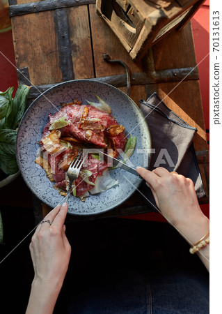 A female hand is preparing a vegetable salad with beef roast beef. A plate of salad with beef roast beef, onions, arugula, pepper and tomatoes on a black table. Top view. 70131613