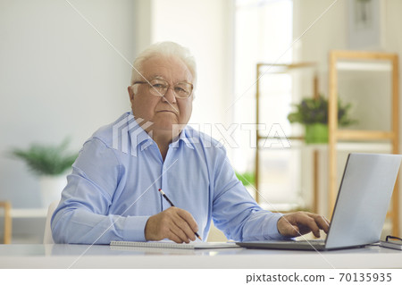 Serious senior man sitting at desk at home using laptop and taking notes in notebook 70135935