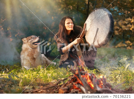 Shaman woman drumming in the sunny forest with an Alaskan Malamute dog 70136221