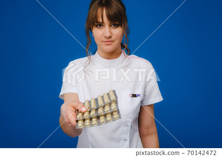 The concept of health care. A young brunette doctor in a white coat on a blue background shows plates with capsules to take. 70142472