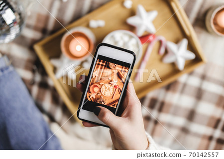 Female hand with smartphone takes pictures of New Year's or Christmas flatlay for social networks. Preparing for holidays, creating content. Wooden tray with cocoa mug marshmallows, candle, stars. 70142557